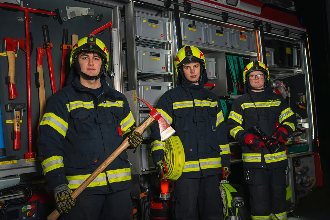 Three firefighters in turnout gear in front of the equipment bay of a fire engine