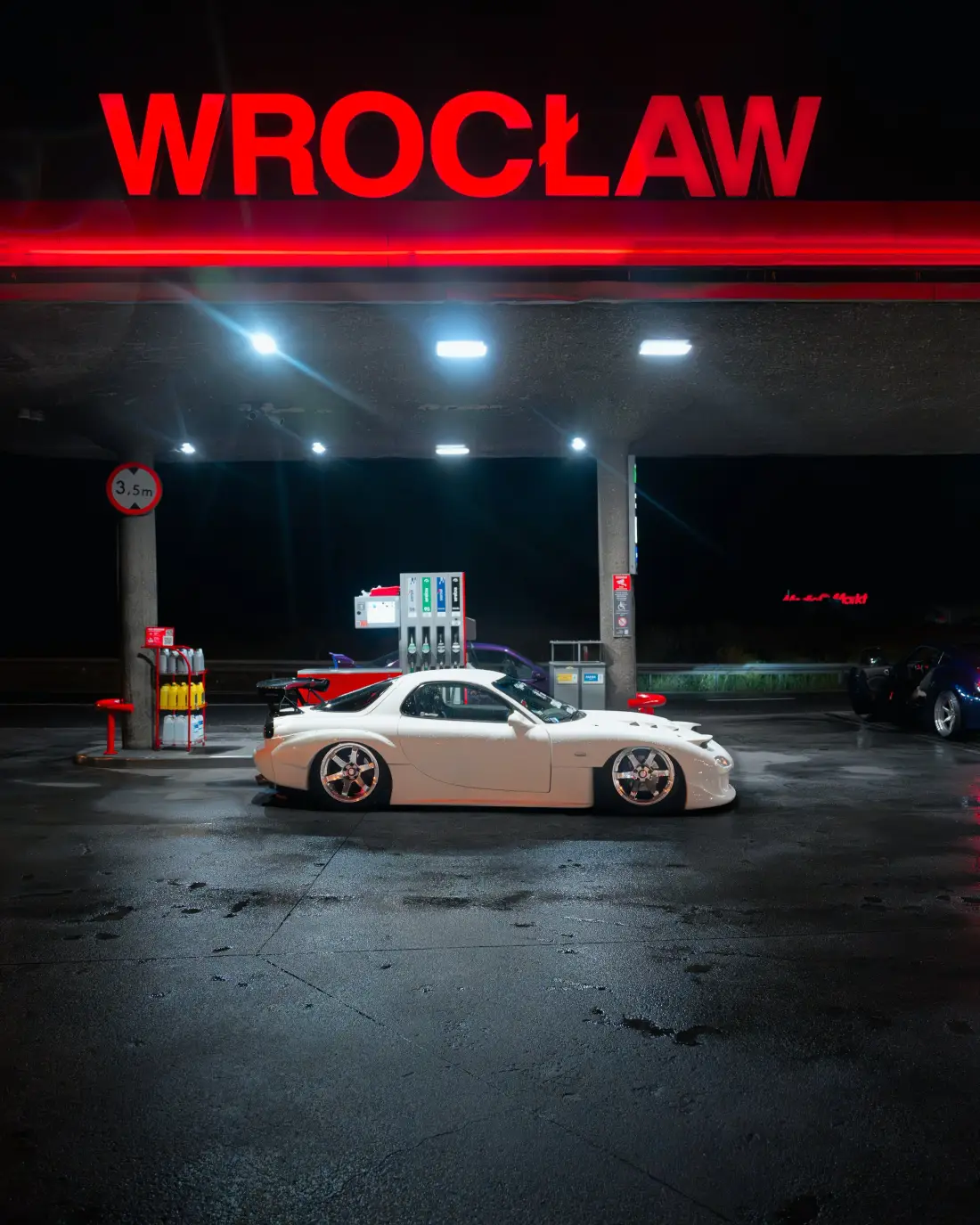 Tankstelle mit rotem Leuchtschriftzug Wrocław bei Nacht, davor ein weißer Sportwagen