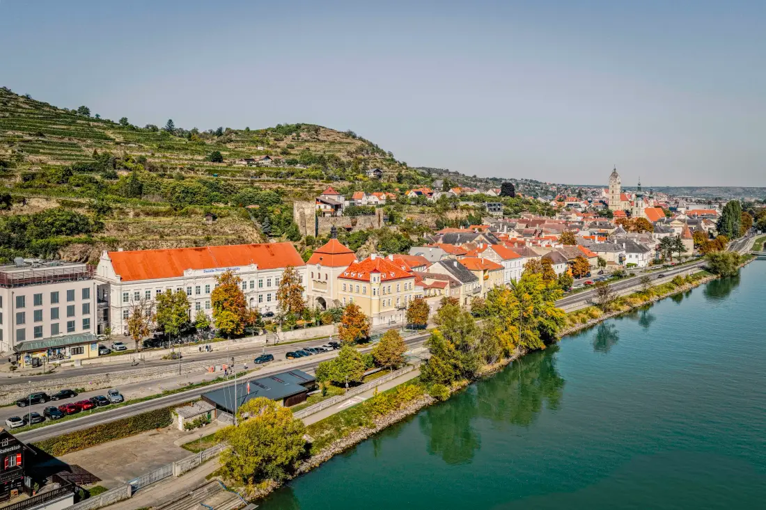 Luftaufnahme einer Stadt am Donauufer, dahinter Weinterrassen