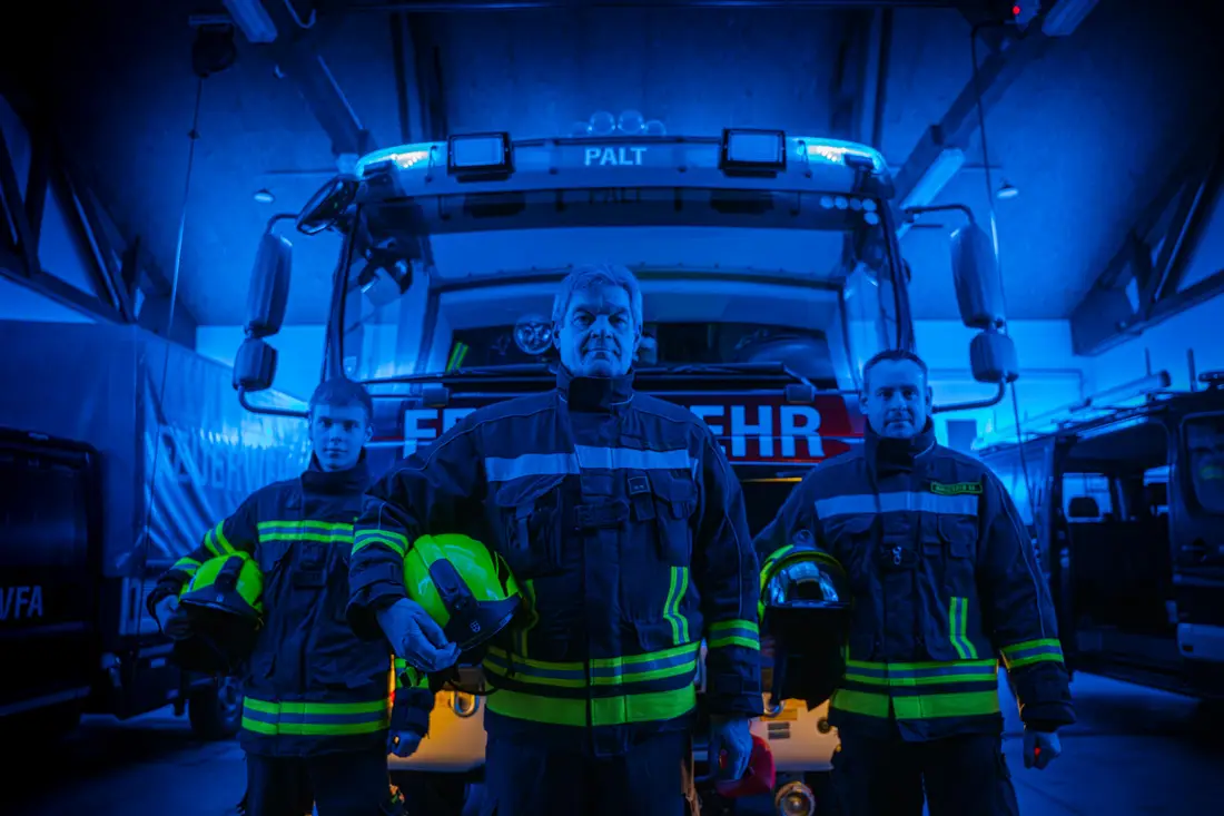 Three firefighters standing in blue light in front of a Palt fire brigade engine