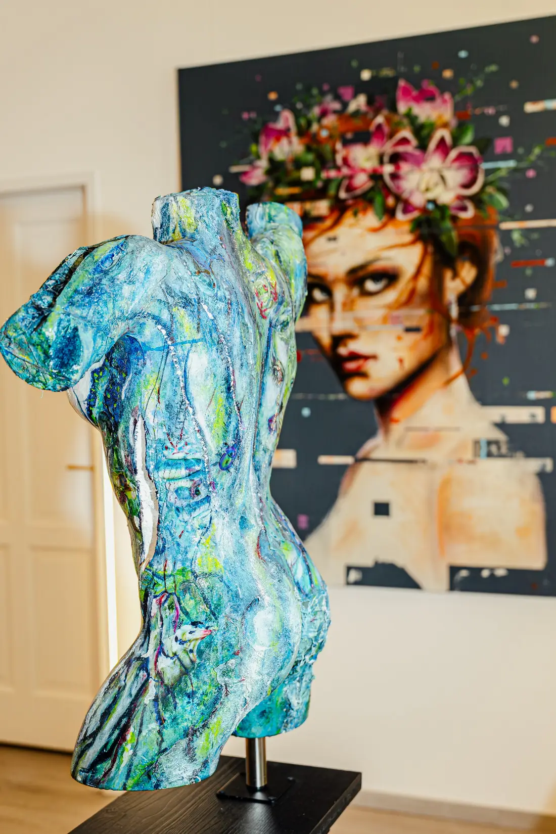 Turquoise painted torso sculpture in front of a portrait painting in a gallery