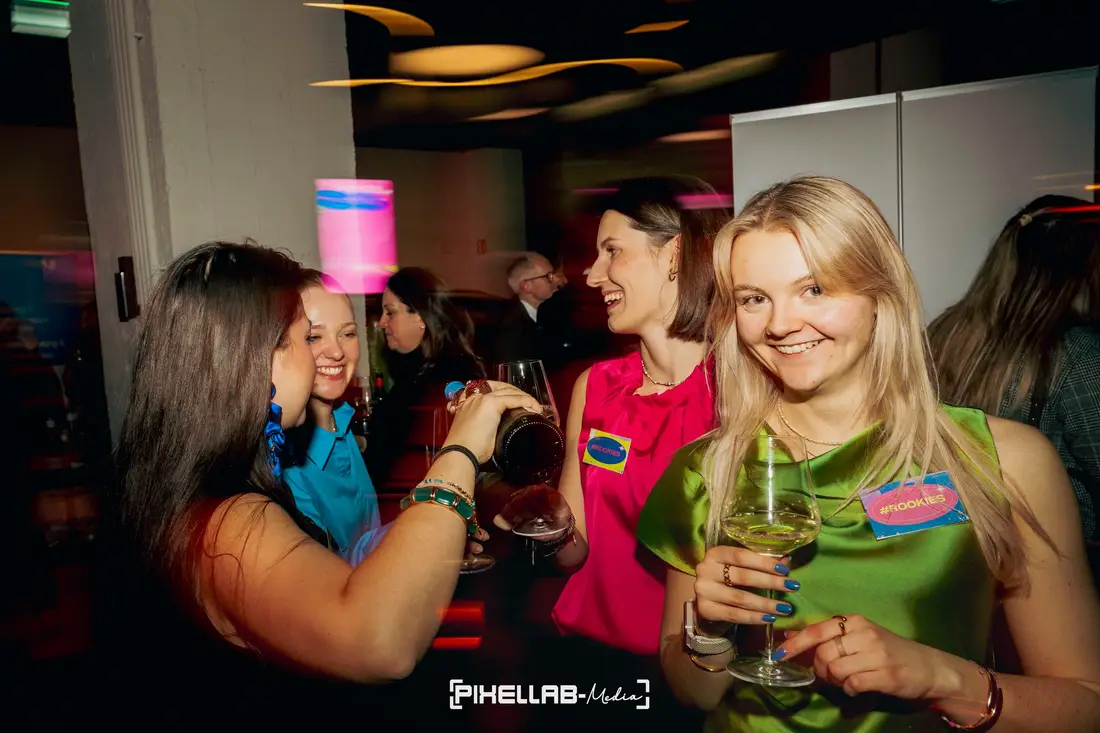 Guests at an evening event: a woman in a green dress smiles at the camera with a wine glass while wine is poured beside her