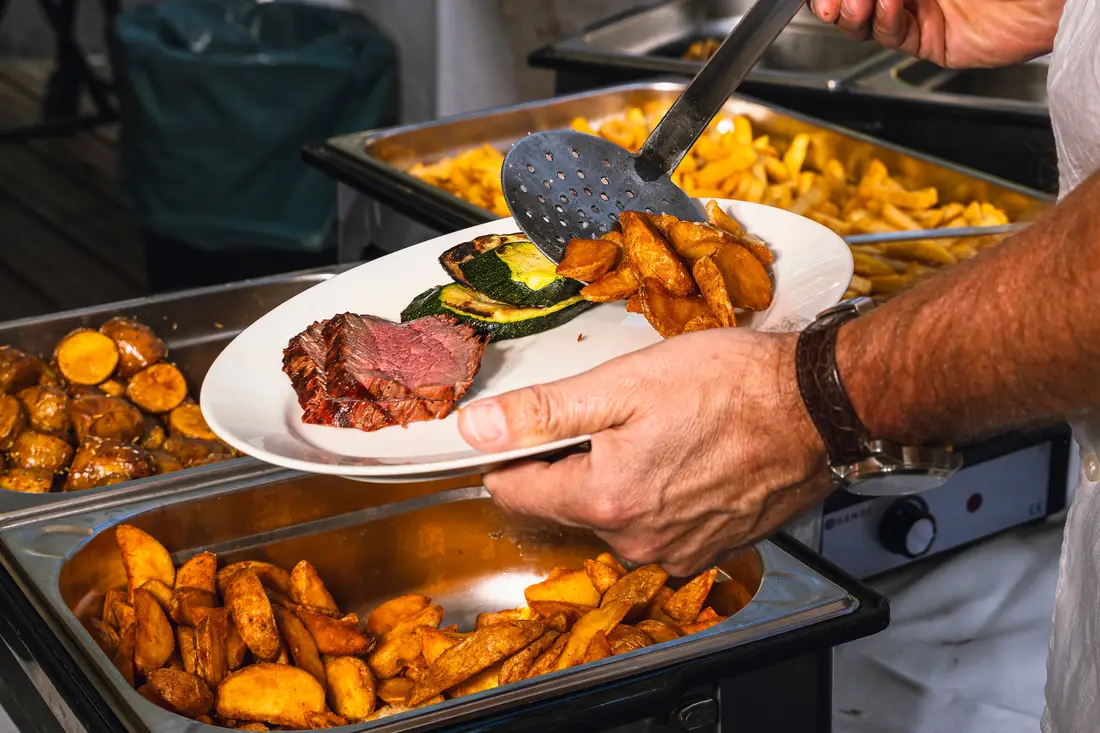 A plate with meat, vegetables and potato wedges being served at a buffet