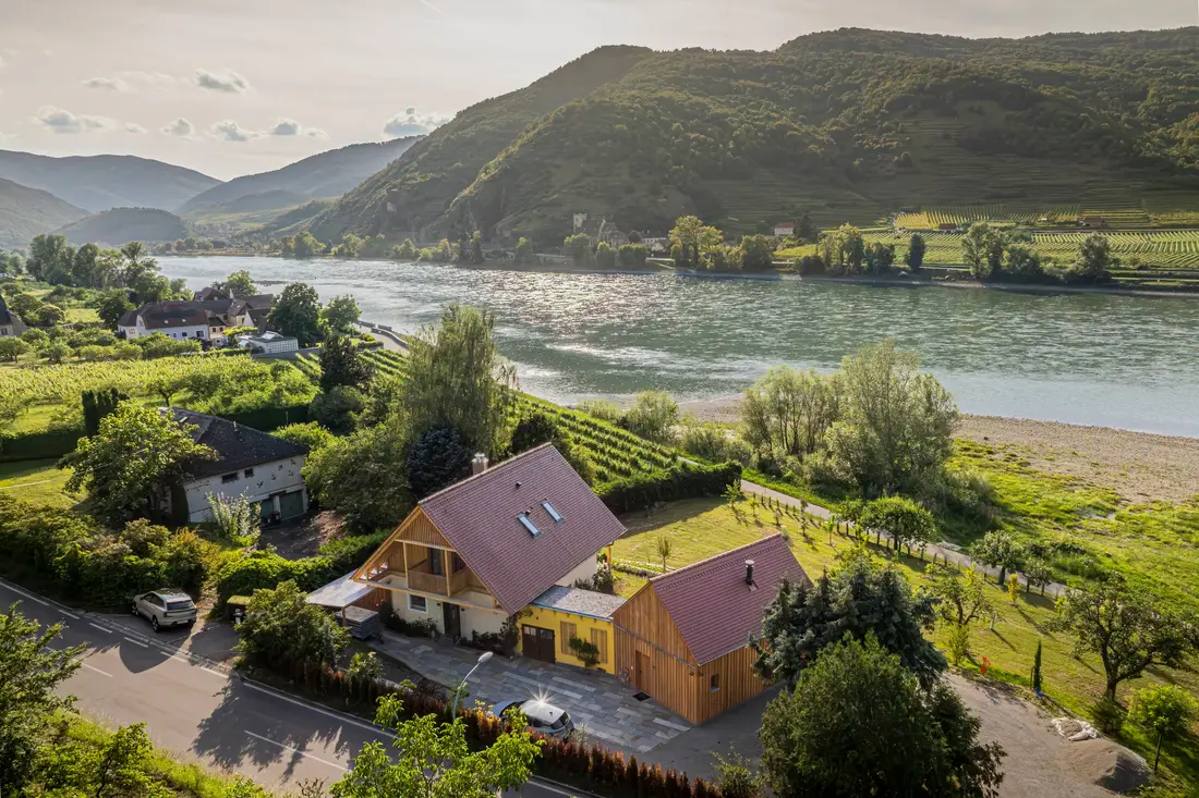 Luftaufnahme von Holzhäusern am Donauufer, dahinter bewaldete Hügel und Weinterrassen