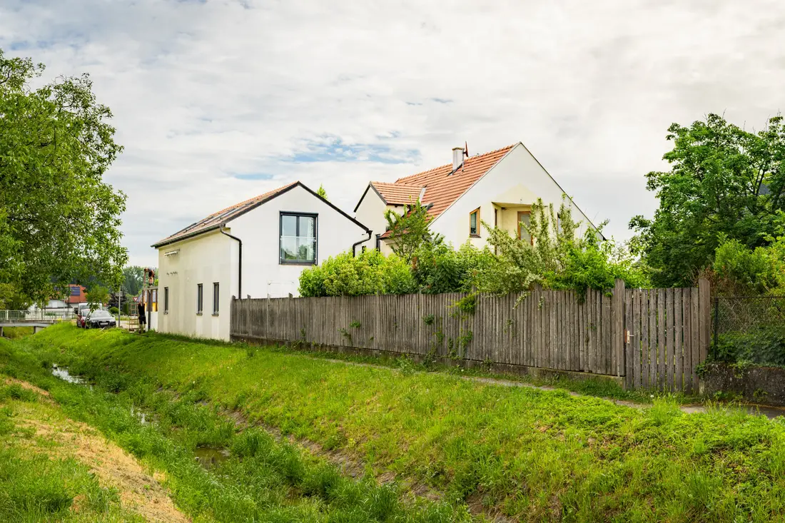 Weißes Wohnhaus mit Garten hinter einem Holzzaun an einem kleinen Bach